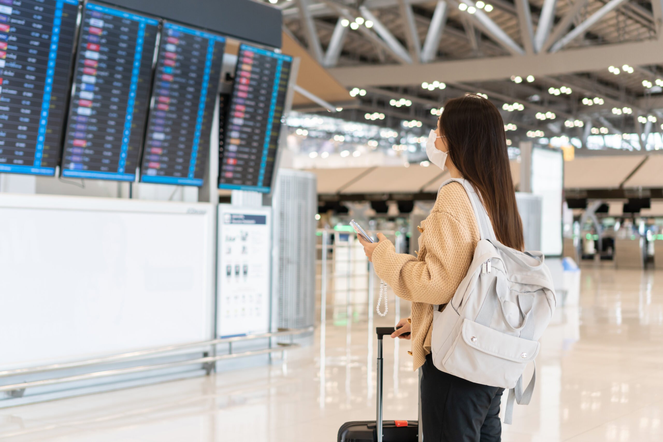 young asian woman wearing medical mask international airport looking flight information board checking her flight travel during coronavirus pandemic new normal lifestyle concept scaled